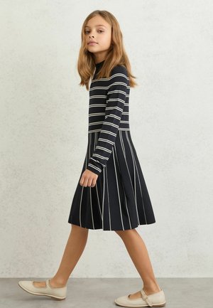 Tüdruk kõnnib, kandes tumesinist kleiti, millel on ülaosas horisontaalsed triibud ja seelikul vertikaalsed triibud, ning sobitatud beežide Mary Jane' stiilis kingadega.