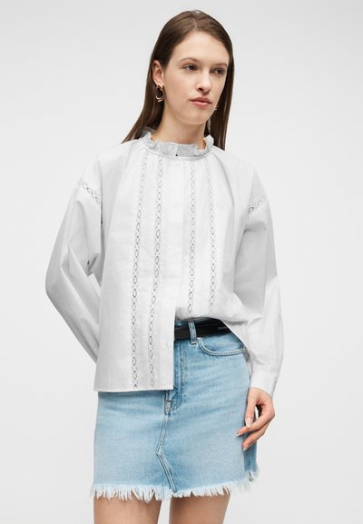 Γυναίκα που φορεί λευκό μπλουζάκι με κάθετες δαντελένιες λεπτομέρειες και γιακά με βολάν, συνδυασμένο με φθαρμένη φούστα από τζιν ανοιχτού μπλε χρώματος και χρυσούς κρίκους σκουλαρίκια.