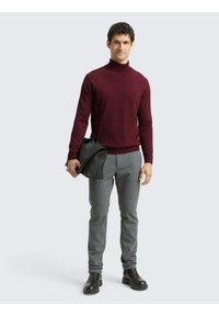 Bordeauxroter Rollkragenpullover, graue maßgeschneiderte Hose und schwarze Stiefeletten. Model hält eine schwarze Tasche und steht vor einem neutralen Hintergrund.
