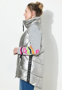 Chaleco inflable plateado con cuello alto, diseño acolchado y acento de logotipo negro. Manga gris con patrón geométrico colorido en los puños.