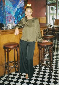 Olivgrön sidenblus med långa ärmar, svarta vida byxor och silversandaler, stående nära en träbar med svart och vit rutigt golv.