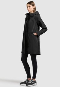 Femme debout portant une longue veste noire à capuche, des leggings noirs et des baskets noir et blanc, une main dans la poche de la veste.