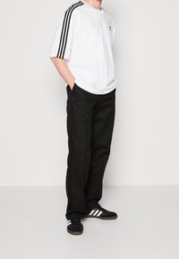 Bílý krátký rukáv tričko s černým trojpruhovým designem na ramenech, spárované s černými kalhotami a černými teniskami se bílými pruhy.