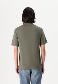 Khaki-grünes Poloshirt mit kurzen Kragen, kurzen Ärmeln und einer glatten Strickqualität. Mit seitlichen Schlitzen und einem leicht verlängerten Rückendesign.