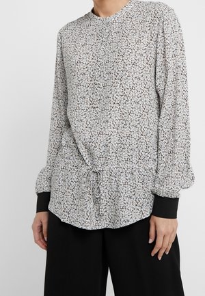 Femme portant un chemisier blanc à manches longues avec un petit imprimé floral noir et un pantalon noir à jambes larges.