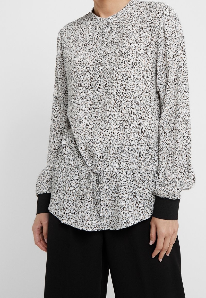 Femme portant un chemisier blanc à manches longues avec un petit imprimé floral noir et un pantalon noir à jambes larges.