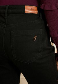 Črne jeans hlače iz denima z oznako iz rjave usnje na pasu, zadnjim žepom z oranžnim vezenjem in ožjim krojem.