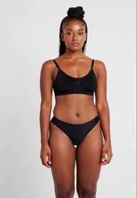 Γυναίκα με πλεξούδα, φορώντας ένα μαύρο σουτιέν και αντίστοιχο μαύρο μπικίνι, στέκεται ενάντια σε απλό ανοιχτό φόντο.