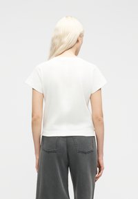 Femme aux cheveux blond platine portant une chemise blanche à manches courtes et un jean gris foncé taille haute, vue de dos.