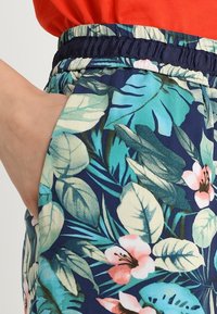 Ręka spoczywa w kieszeni granatowych spodni z elastycznym pasem, ozdobionych niebiesko-zielonymi liśćmi i różowym kwiatowym wzorem, ubrana w pomarańczową bluzkę.