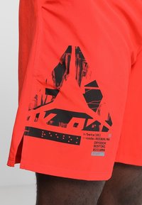 Shorts de sport orange vif avec logo abstrait noir et texte, portés par une personne, avec une fente latérale près de l’ourlet.