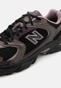 Svart och lila New Balance träningsskor med nätpaneler, vadderad krage, svarta snören och strukturerad gummisula på vit bakgrund.
