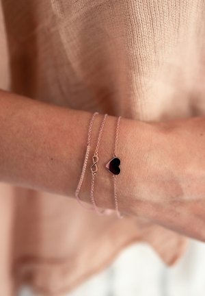 Dos pulseras de oro rosa en una muñeca: una presenta una cadena rosa con un símbolo de infinito, la otra tiene un colgante negro en forma de corazón.