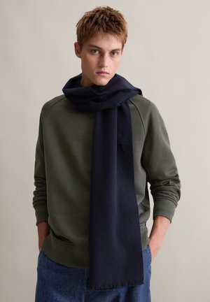 Navy wool scarf draped casually over a green sweatshirt, featuring a soft texture and fringed ends. Simple design without visible patterns.