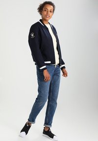 Marine bomberjakke med hvite striper, prydet med broderte lapper; kombinert med en lys beige topp og mid-blue jeans, svarte slip-on sko.
