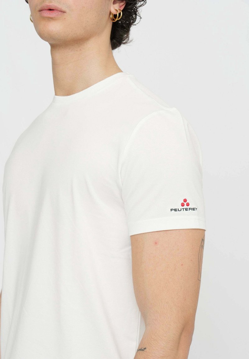 Jeune personne portant un t-shirt blanc uni à manches courtes avec un logo Peuterey rouge et noir sur la manche gauche.