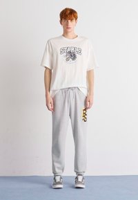 Hvid grafisk t-shirt med "STAPLE" logo, parret med lysegrå sweatpants med gule accenter og et broderet design.