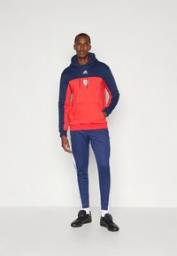 Sweat à capuche Adidas avec un corps rouge et des manches bleu marine, poche kangourou, capuche à cordon, porté avec un pantalon de survêtement bleu et des baskets noires.