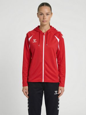 Femme debout, face à l'avant, portant un sweat à capuche rouge zippé avec des accents blancs et un pantalon noir avec des bandes chevrons blanches.