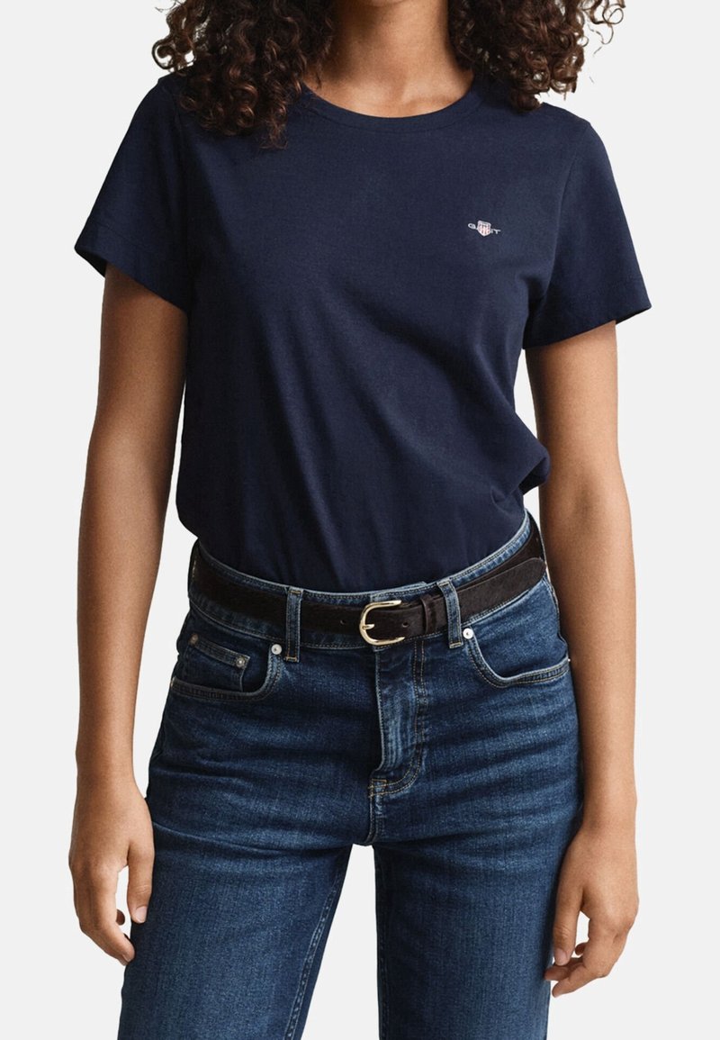 Femme portant un t-shirt bleu marine à manches courtes rentré dans un jean bleu foncé avec une ceinture noire et une boucle dorée, debout, bras détendus.