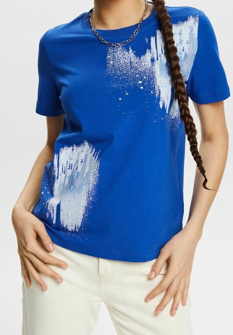 Chemise bleue à manches courtes avec motif éclaboussures de peinture blanche, portée avec un pantalon clair, un collier chaîne argenté et de longs cheveux tressés sur l'épaule.