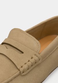 Loafer από ανοιχτό καφέ καστόρι με στρογγυλεμένη μύτη, ραμμένες λεπτομέρειες και διακοσμητικό λουρί πάνω από το μπροστινό μέρος. Η εσωτερική επένδυση είναι καμηλό.