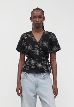 Femme portant un chemisier cache-cœur noir avec une broderie florale blanche et un jean taille haute bleu clair, debout devant un fond blanc uni.