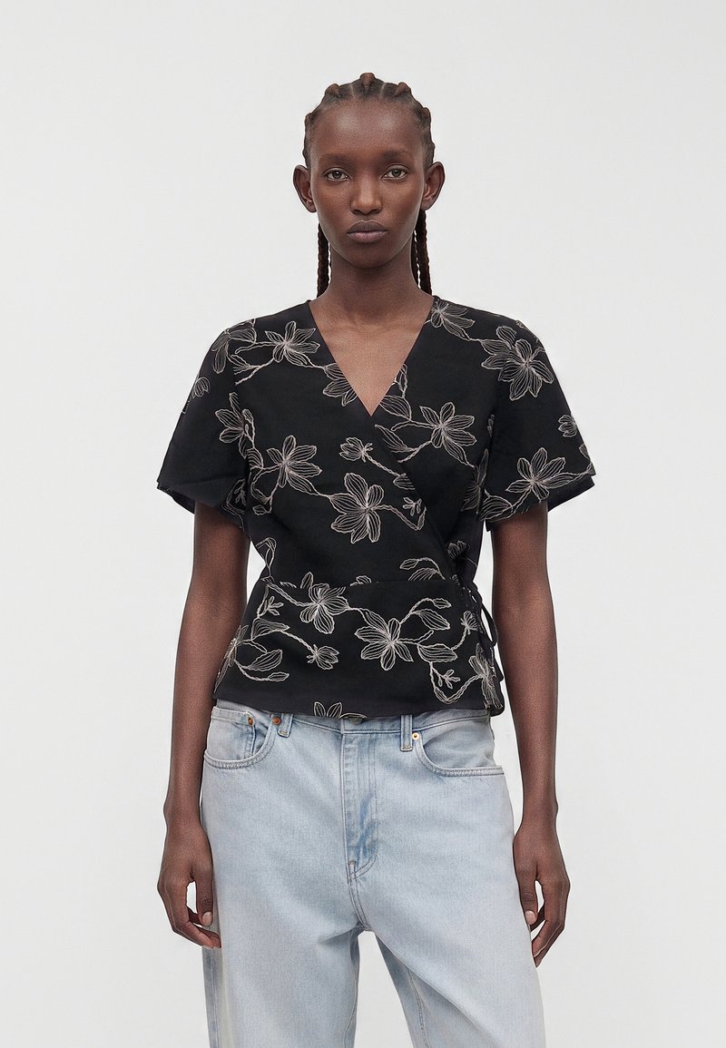 Femme portant un chemisier cache-cœur noir avec une broderie florale blanche et un jean taille haute bleu clair, debout devant un fond blanc uni.