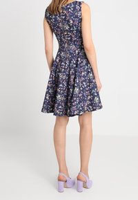 Marinblå blommig klänning med passform som är figursydd upptill och utsvängd nertill, ärmlös design, dragkedja bak och en utsvängd kjol. Bärs med lavendelfärgade klackskor.