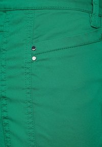 Grön denimtyg med en närbild av en ficka, med synliga sömmar och två silverfärgad knappdetaljer. Slät textur med minimal glans.