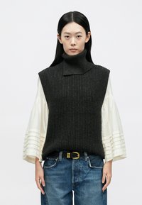 Donkergrijze gebreide mouwloze coltrui met een gestructureerd ribpatroon, gecombineerd met een wit geplooid shirt met lange mouwen en een blauwe spijkerbroek.