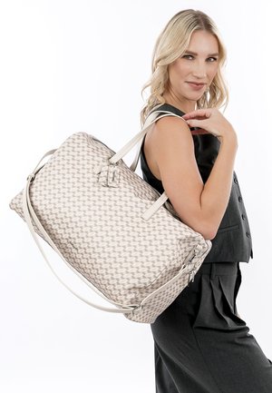Femme blonde portant un haut sans manches foncé et un pantalon, portant un grand sac fourre-tout beige à motifs sur l'épaule, souriant à la caméra.