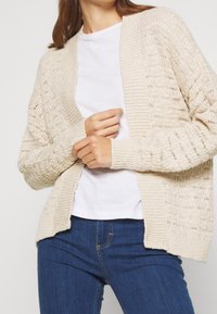 Beige stickad cardigan med öppen framsida, prydd med strukturerade stickmönster och ribbade detaljer på ärmar och nederkant. Kombinerad med en vit topp och blå jeans.