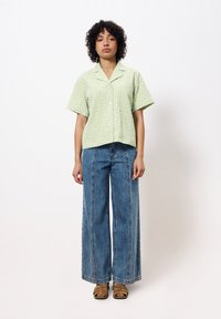 Grün-weiß kariertes kurzärmeliges Hemd, weit geschnittene blaue Jeans mit Mittelnaht und braune Riemchensandalen. Einfarbiger Hintergrund.