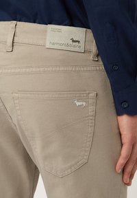 Beige katoenen broek met een achterzak, voorzien van lichte stiksels en een klein geborduurd hondenlogo. Etiket zichtbaar op de tailleband.