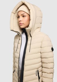 Giacca puffer beige con un ampio cappuccio, chiusura a zip e tasche laterali. Presenta cuciture orizzontali e un accento nero sul manica.