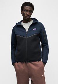 Tumši zils un melns rāvējslēdzēja džemperis ar kapuci un pelēkām detaļām, priekšpusē uz krūtīm ar violetu Nike logotipu un sānu rāvējslēdzēja kabatu.