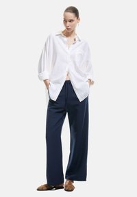 Witte linnen blouse met knopen, marineblauwe wijde broek, bruine platte sandalen. De blouse heeft opgerolde mouwen en een borstzak; de broek heeft een trekkoord.