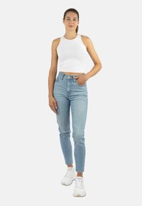 Frau steht, trägt ein weißes ärmelloses Crop Top, hellblaue High-Waist-Jeans, weiße Sneakers und gelbe Ohrringe, vor einem schlichten weißen Hintergrund.