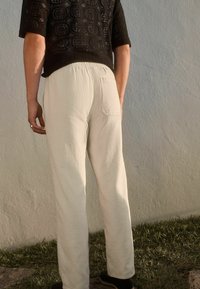 Pantalon en lin beige clair avec une coupe décontractée et une poche arrière, associé à un haut en tricot noir texturé. Sur un fond neutre.