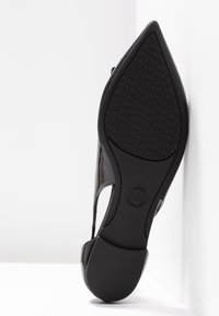Semelle d'une chaussure noire à bout pointu avec motif texturé et logo MK sur la bande de roulement, photographiée sur fond blanc.