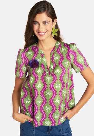 Woman wearing green and pink patterned short-sleeve blouse with floral brooch and green leaf-shaped earrings, smiling with hands in pockets.