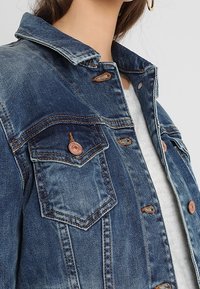 Denim jacket featuring a classic button-up design, light blue color, copper-toned buttons, and chest pockets with detailed stitching.