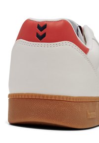 White leather sneaker with red heel accent, featuring black chevron design. Gum rubber sole with textured finish and triangular pattern.