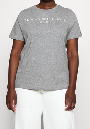 Person wearing a gray Tommy Hilfiger t-shirt with white text and logo centered on the chest, paired with white pants.