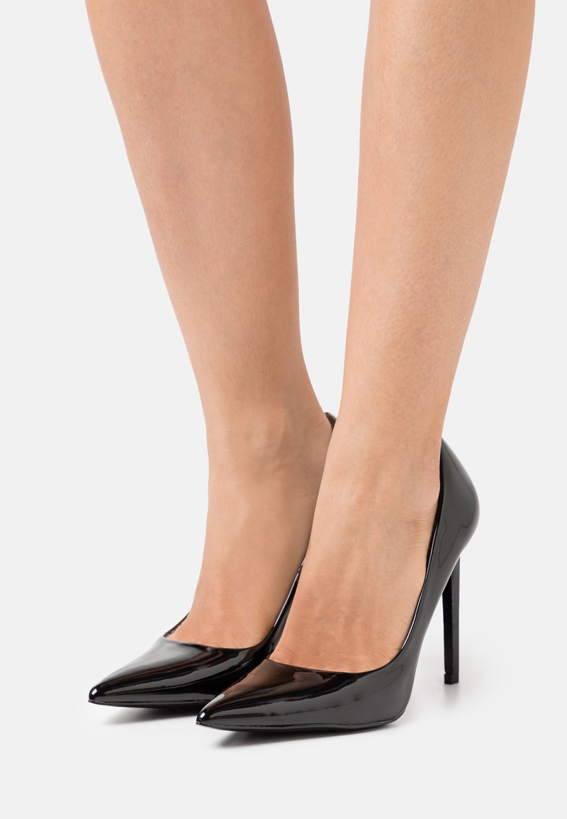 Jambes portant des escarpins noirs en cuir verni à bout pointu avec talons aiguilles sur un fond blanc uni.