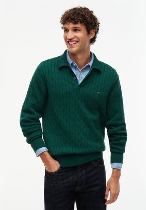 Jeune homme souriant, portant un pull torsadé vert foncé par-dessus une chemise en denim bleu clair avec un jean foncé, debout avec une main dans la poche.