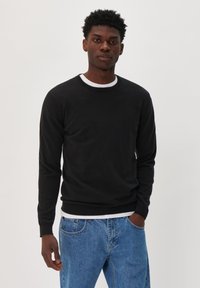 Black crew neck sweater with long sleeves, made of a soft material. Features a ribbed hem and cuffs, worn over a white shirt and paired with blue jeans.