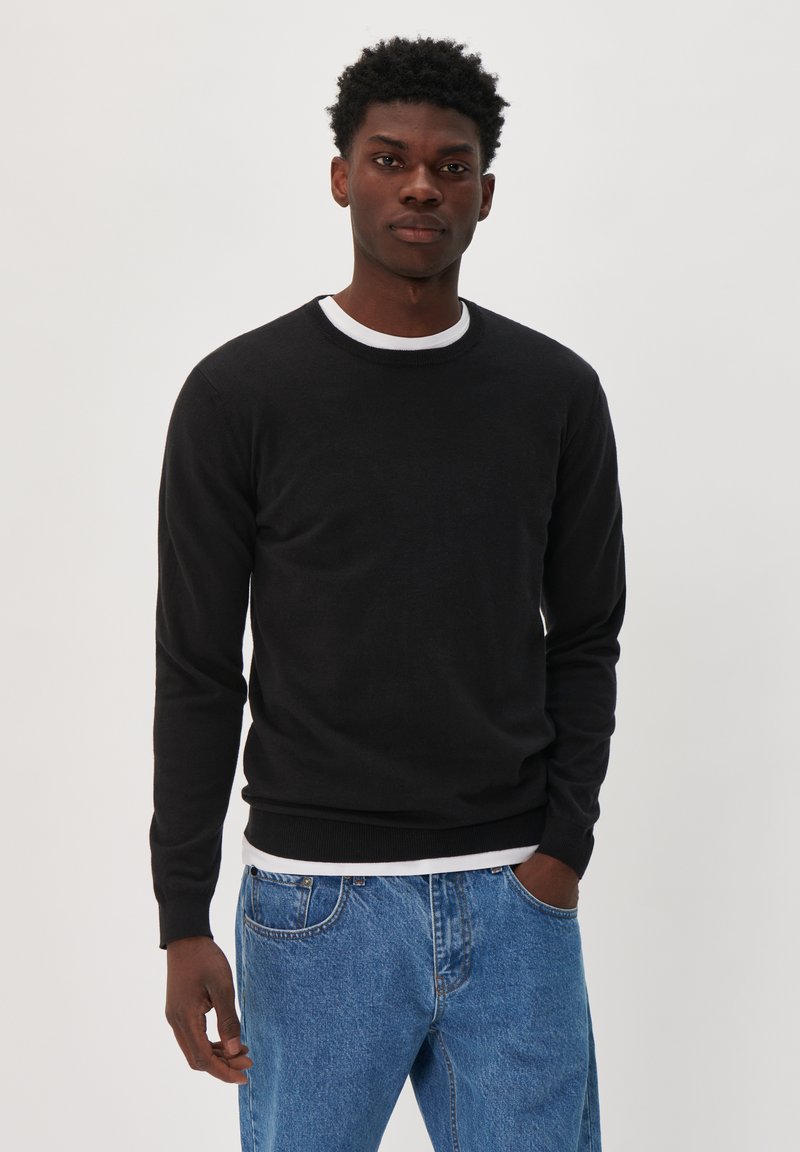 Black crew neck sweater with long sleeves, made of a soft material. Features a ribbed hem and cuffs, worn over a white shirt and paired with blue jeans.