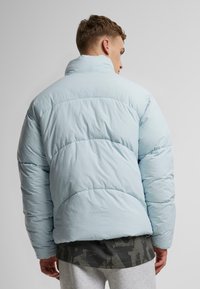 Ljusblå vadderad jacka med hög krage, kviltat mönster och elastiska manschetter. Lager på en mörkt mönstrad skjorta och grå sweatpants.